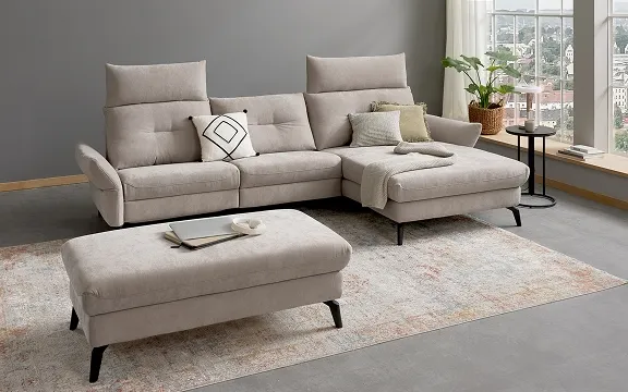 Moderner, hellgrauer Eck-Sofa-Zweiteiler mit passendem Hocker auf einem Teppich vor grauer Wand.